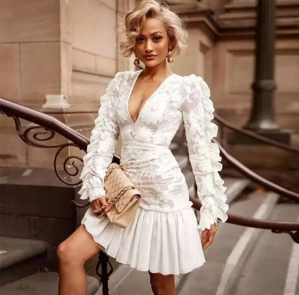 Robe courte blanche à manches longues plissée col V portée par une femme élégante en milieu urbain.